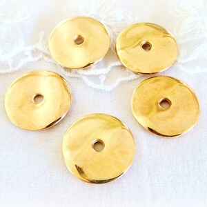 May include: Five gold-coloured metal round disc beads with a hole in the centre. The beads are on a white fabric surface.
