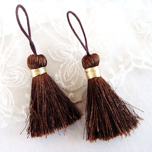 May include: Two brown tassels with gold trim. The tassels are made of soft, fluffy fibers and have a loop at the top for hanging.