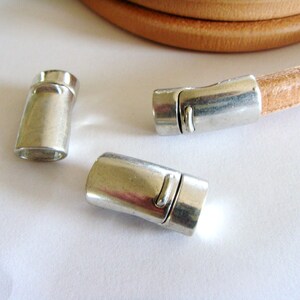 May include: Close-up of silver magnetic clasps and a tan leather cord. The clasps are cylindrical with a rectangular opening. The leather cord is coiled in the background. Ideal for jewelry making.