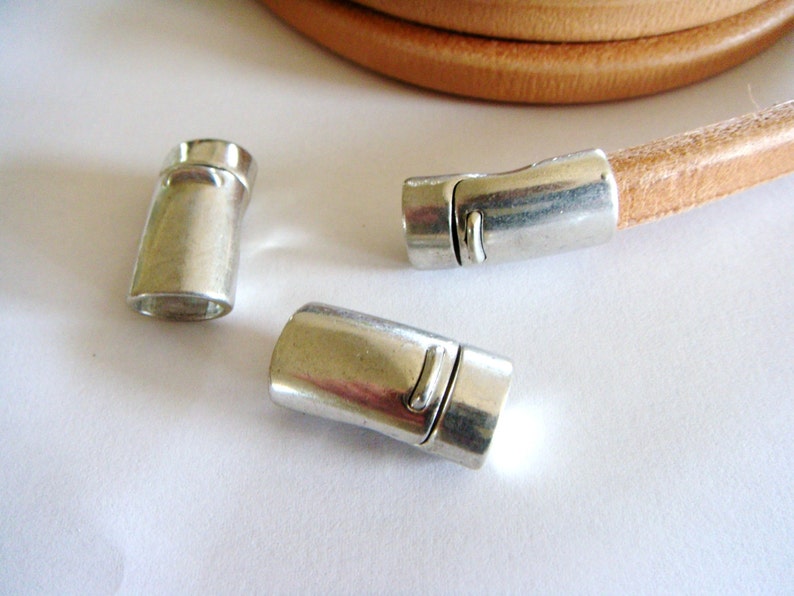 May include: Close-up of silver magnetic clasps and a tan leather cord. The clasps are cylindrical with a rectangular opening. The leather cord is coiled in the background. Ideal for jewelry making.