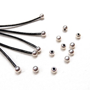 Puede incluir: Primer plano de cuentas plateadas y cordones de cuero negro. Las cuentas son redondas y tienen un acabado liso y metálico. Los cordones son finos y flexibles.