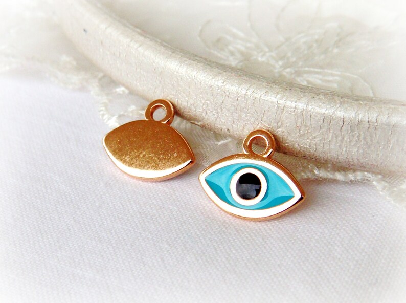Puede incluir: Dos abalorios de metal dorado en forma de ojo. Un abalorio es una forma de ojo s&oacute;lida, mientras que el otro abalorio tiene un ojo de esmalte azul y blanco con una pupila negra.