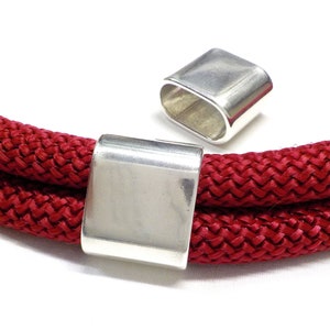 May include: Two silver metal cord end caps for a red rope or cord. The cord end caps are rectangular in shape and have a smooth, polished finish.