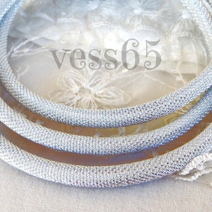 May include: Three silver-colored, mesh-like necklaces are arranged in a circular pattern. The necklaces are made of a shiny, metallic material. The text "vess65" is visible in the background.