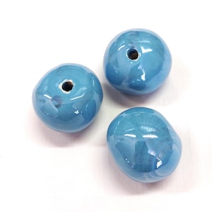 Könnte beinhalten: Drei glänzende, hellblaue Keramikperlen mit unregelmäßigen Formen und einem Loch in der Mitte. Die Perlen haben eine glatte, reflektierende Oberfläche und sind vor einem weißen Hintergrund angeordnet.