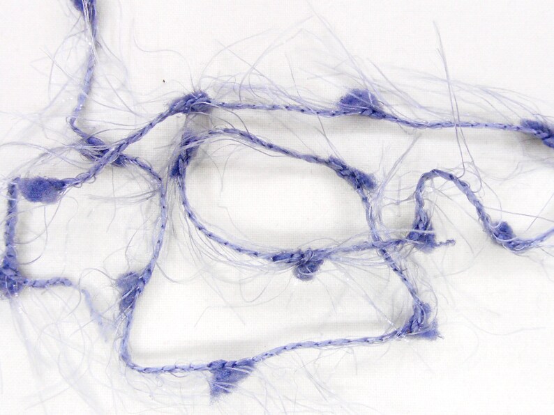 May include: Close-up of a tangled, light purple yarn with a fuzzy texture. The yarn is composed of thin, transparent strands and thicker, knotted sections of a darker purple color. The background is plain white, highlighting the yarn's intricate details.