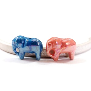 Op de afbeelding: Twee keramische olifantbedels, één blauw en één roze, met iriserende afwerkingen. De bedels liggen op een wit oppervlak.