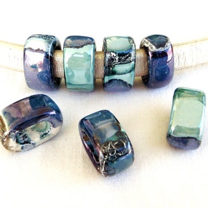 May include: Five ceramic beads in shades of blue and green with iridescent accents. The beads are rectangular and have a hole in the center. They are strung on a white leather cord.