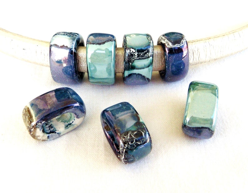 May include: Five ceramic beads in shades of blue and green with iridescent accents. The beads are rectangular and have a hole in the center. They are strung on a white leather cord.