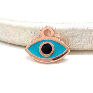 Puede incluir: Un amuleto de ojo turco de oro rosa con un ojo azul turquesa y una pupila negra. El amuleto tiene un bucle en la parte superior para sujetarlo a un collar o pulsera.