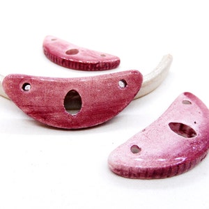 May include: Three pink ceramic crescent moon shapes with holes. The moon shapes are on a white surface.
