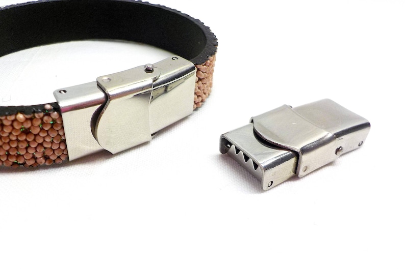 May include: A close-up of a silver metal clasp for a bracelet. The clasp is made of two pieces that snap together. The clasp is shown against a white background.