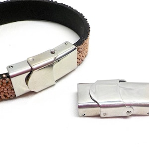 May include: A close-up of a silver metal bracelet clasp. The clasp is made of two pieces that fit together to secure the bracelet. The bracelet is made of brown leather with small, round beads.