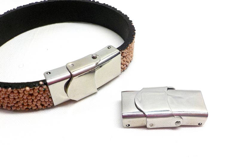May include: A close-up of a silver metal bracelet clasp. The clasp is made of two pieces that fit together to secure the bracelet. The bracelet is made of brown leather with small, round beads.
