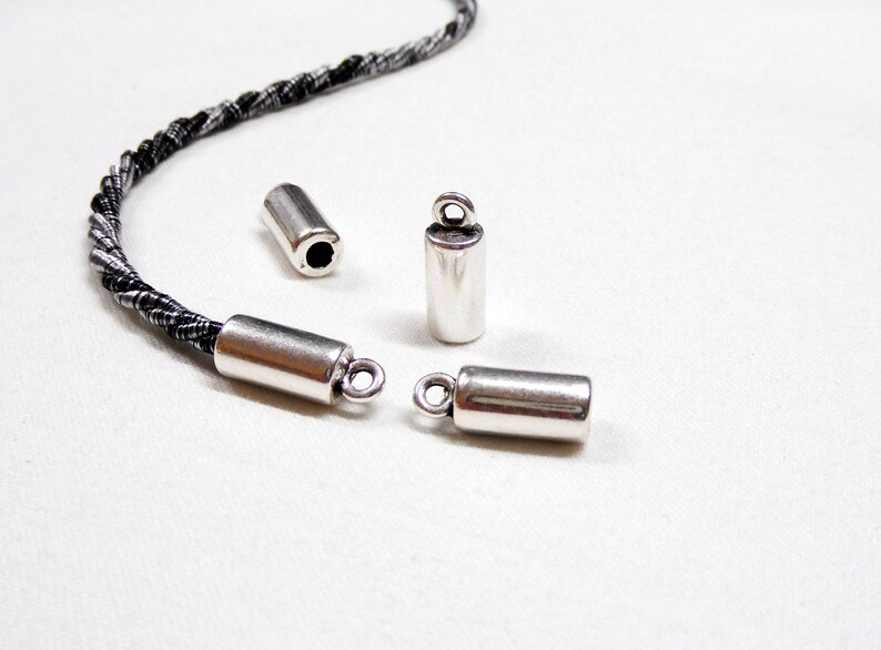 May include: Four silver metal cord end caps. Two of the caps have a loop on the end. The caps are on a white surface.
