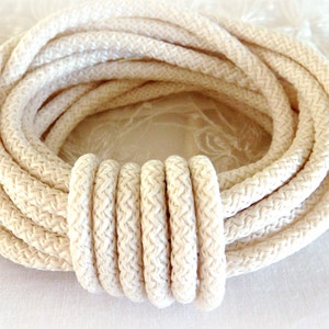 Pode incluir: Corda creme enrolada com um padrão tecido texturizado. A corda é disposta em forma circular, com uma seção da corda agrupada. A corda é de cor neutra, adequada para vários projetos de artesanato.
