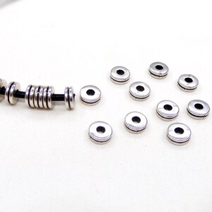 Puede incluir: Cuentas espaciadoras de metal plateado para la elaboración de joyas. Las cuentas son redondas con un pequeño agujero en el centro. Hay 12 cuentas en total, algunas están en un cordón de cuero negro.