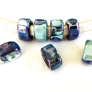 May include: A set of six rectangular ceramic beads with a blue, green, and silver glaze. The beads are strung on a white leather cord.