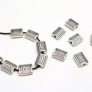 May include: Silver-colored rectangular beads with a recessed geometric design. Some beads are strung on a black cord, while others are loose. The beads are approximately 0.5 inches long.