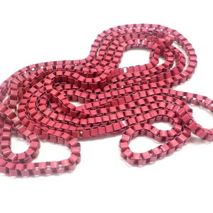 Puede incluir: Una cadena larga de eslabones metálicos rojos, dispuestos en un patrón suelto y fluido. La cadena está hecha de pequeños eslabones cuadrados.