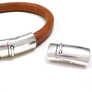 May include: A brown leather bracelet with a silver magnetic clasp. The clasp is cylindrical with a curved design and a small loop. The bracelet is shown with the clasp open and closed.