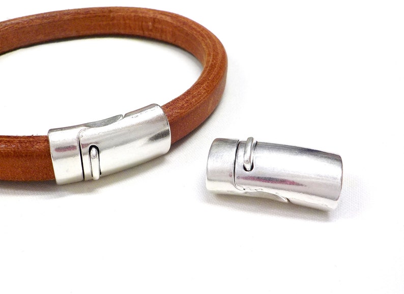 May include: A brown leather bracelet with a silver magnetic clasp. The clasp is cylindrical with a curved design and a small loop. The bracelet is shown with the clasp open and closed.