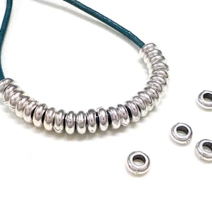 May include: A necklace with silver-coloured, rounded beads on a teal cord. Several loose beads are scattered nearby. The beads have a shiny, metallic finish, and the cord is visible at the top.