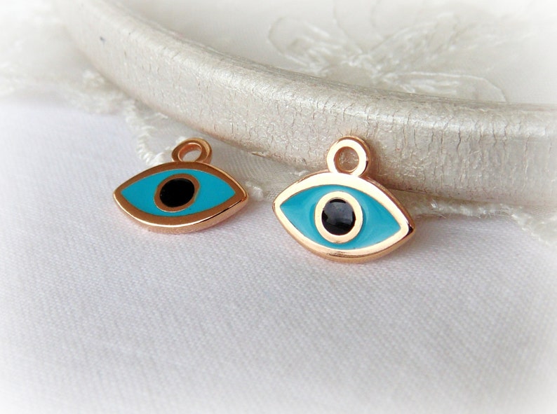 Puede incluir: Dos amuletos de ojo turco dorados con esmalte azul y centros negros. Los amuletos tienen un bucle en la parte superior para sujetarlos a joyas.