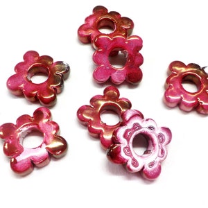Puede incluir: Seis cuentas de color rosa y blanco con forma de flor y un agujero en el centro. Las cuentas tienen un acabado brillante.