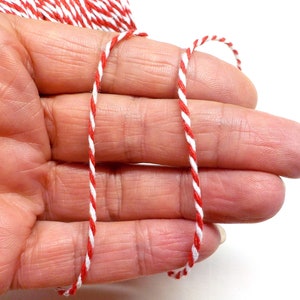 May include: Red and white baker's twine, a type of string used for wrapping and tying, is shown in a hand. The twine is twisted and has a striped pattern.