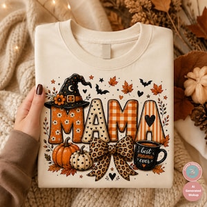 Può includere: Una felpa color crema con una stampa grafica della parola MAMA in motivi arancioni e bianchi. Il design include un cappello da strega, zucche, un fiocco con stampa leopardata e una tazza nera con il testo best mama ever, circondato da foglie autunnali.