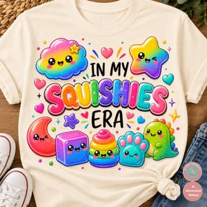 Op de afbeelding: Een cr&egrave;mekleurig t-shirt met een kleurrijke afbeelding en de tekst "IN MY SQUISHIES ERA". Het ontwerp bevat een regenboogwolk, een regenboogster, een rode maansikkel, een paarse kubus, een regenboogstapel, een blauwe poot en een groene dinosaurus.