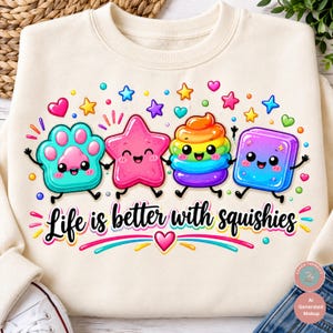 Op de afbeelding: Een cr&egrave;mekleurig sweatshirt met een kleurrijke afbeelding van vier stripfiguren: een turquoise poot, een roze ster, een regenboogwerveling en een blauwe kubus. Onder de figuren staat de tekst "Life is better with squishies" in zwarte letters.