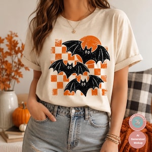 Op de afbeelding: Een crèmekleurig T-shirt met korte mouwen en een grafische print van drie zwarte vleermuizen die voor een oranje zon vliegen, met een verweerd oranje en wit geblokt patroon. Het shirt heeft een ronde hals en opgerolde mouwen.