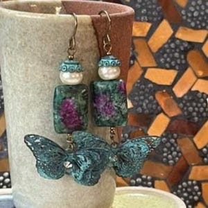 May include: Dangle earrings featuring a dark teal butterfly charm with a textured finish, a rectangular green and magenta stone bead, and a white spherical bead. The earrings are suspended from bronze-toned metal hooks and display a layered design.