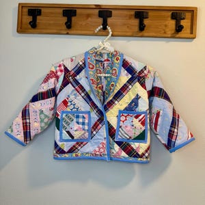 May include: A patchwork jacket with a variety of colorful fabric patterns, including plaid, floral, and geometric designs. The jacket has a blue trim, two front pockets, and is hanging on a wooden rack with black hooks.