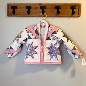 Puede incluir: Chaqueta acolchada con un estampado de estrellas en tonos rosas y morados, con detalles blancos y florales. La chaqueta está colgada en un perchero de madera con ganchos negros. La chaqueta tiene cierre de lazo y una etiqueta.