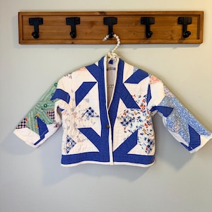 May include: A handmade quilted jacket in white, blue, and floral patterns hangs on a wooden rack. The jacket features a geometric design with blue triangles and floral patches. The jacket is a unique piece of wearable art.