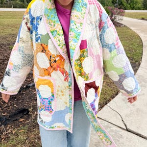 May include: A patchwork jacket with a variety of colorful fabric patterns, including floral and geometric designs. The jacket has a light pink trim and two front pockets. The sleeves are long and the jacket is open in the front.