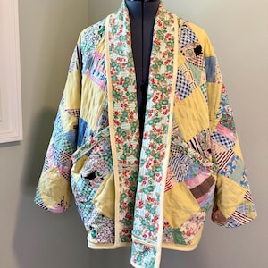 Könnte beinhalten: Patchwork-Steppjacke mit gelber Basis und verschiedenen bunten Stoffquadraten. Die Jacke hat einen Blumendruck auf den Revers und den Besätzen, mit einem cremefarbenen Rand. Die Jacke wird auf einer Schaufensterpuppe präsentiert.