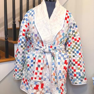 May include: A quilted robe with a patchwork design in red, blue, and white squares. The robe has a white shawl collar and a matching belt. The robe is displayed on a mannequin.