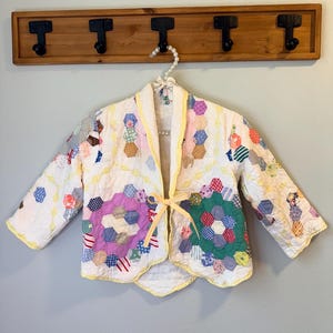 May include: A white quilted jacket with a patchwork design featuring various colors and patterns. The jacket has a yellow trim and a yellow ribbon closure. It is hanging on a wooden rack with black hooks.
