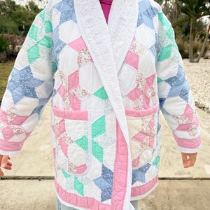 May include: A quilted jacket featuring a patchwork design in white, pink, blue, and green. The jacket has a white collar and trim, with two front pockets. The pattern includes floral and geometric shapes, creating a vintage aesthetic.