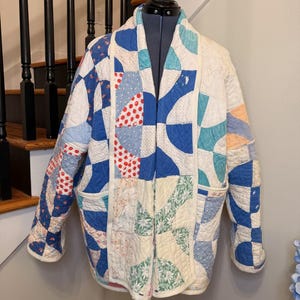 May include: A quilted jacket with a patchwork design in shades of blue, white, and red. The jacket features a variety of patterns, including polka dots, floral prints, and geometric shapes. It has a relaxed fit with pockets and a shawl collar.