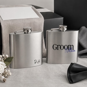 May include: Two brushed stainless steel hip flasks with screw-top caps. One flask features the black text "D.H" in the bottom corner. The other flask displays the black text "Groom" with the blue script name "Nathan" printed underneath.
