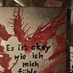 Könnte beinhalten: Eine goldene Leinwand mit roten Farbstichen und dem Text "Es ist okay wie ich mich fühle." in schwarzer Farbe.