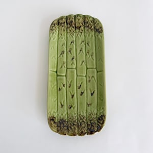 May include: A green ceramic dish shaped like asparagus. The dish has a brown glaze on the bottom and a green glaze on the top.