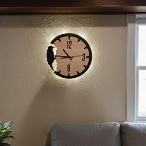 Könnte beinhalten: Eine runde Wanduhr aus Holz mit schwarzem Rand und einer Vogelsilhouette. Das Zifferblatt hat schwarze Ziffern bei 12, 3, 6 und 9 Uhr. Die Uhr ist an einer beigefarbenen Wand befestigt.