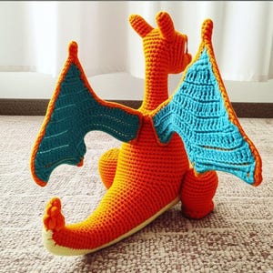 May include: A handmade crochet dragon in orange, teal, and yellow. The dragon has large wings with teal and orange detailing. The dragon's body is orange, with a long tail and small feet. The dragon is sitting on a neutral-colored surface.