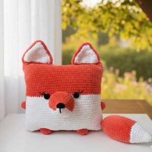 May include: A handmade crochet fox pillow in orange and white. The square pillow features a fox face with a round nose, black eyes, and small ears. A matching orange and white tail is next to the pillow.